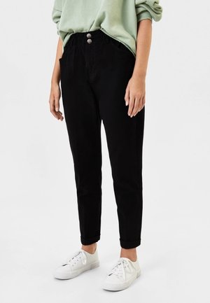 Personne portant un pantalon noir taille haute avec les revers roulés, un haut vert clair à manches longues et des baskets blanches sur fond blanc.