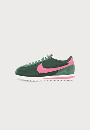 Baskets basses vertes avec virgule Nike rose et languette au talon, lacets blancs et semelle blanche avec bande rose sur fond uni.
