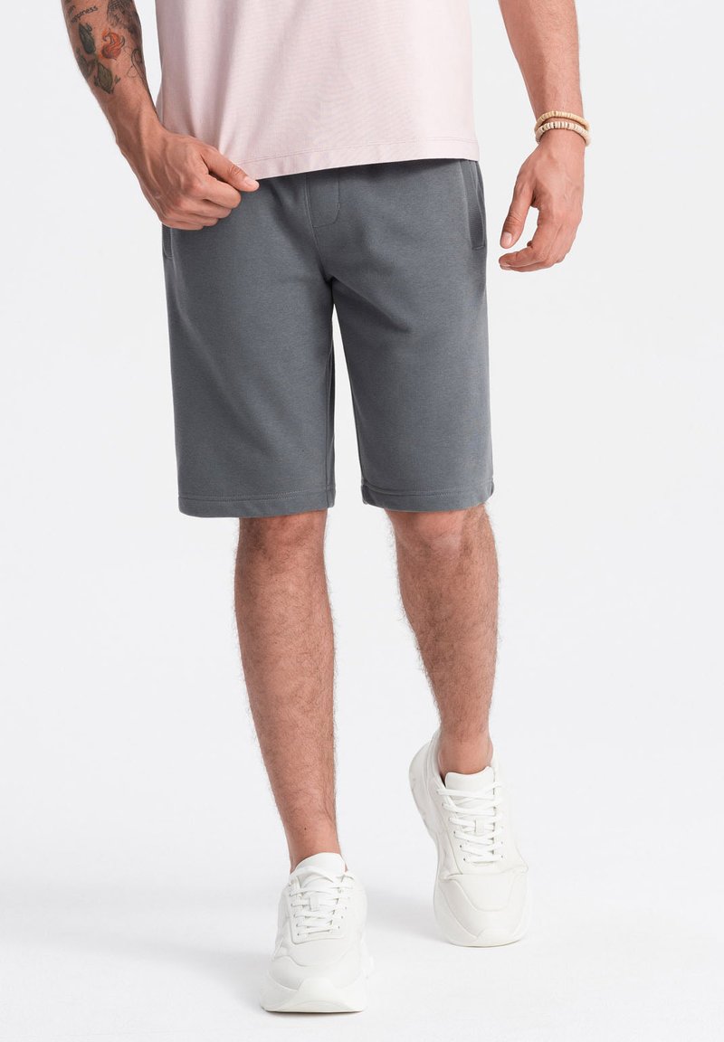 Graue Shorts aus weichem Stoff mit elastischem Bund, seitlichen Taschen und knielangem Schnitt, kombiniert mit weißen Turnschuhen.