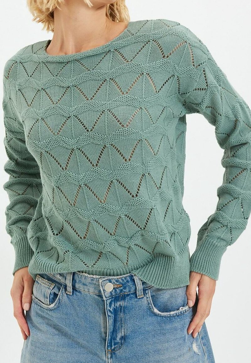 Pull vert clair en maille avec motif géométrique ajouré, porté avec un jean denim bleu clair, montrant la taille et les mains.