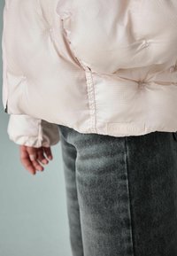 Dikke lichtroze jas met een gestructureerde afwerking, voorzien van elastische manchetten en een label. Draagt met donkergrijze gestructureerde denim broek.