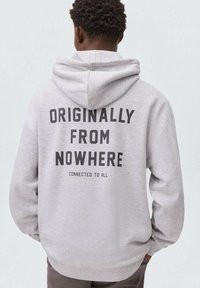 Sweatshirt gris avec une poche kangourou et un grand texte imprimé au dos, indiquant "ORIGINAIRES DE NULLE PART CONNECTÉS À TOUS."