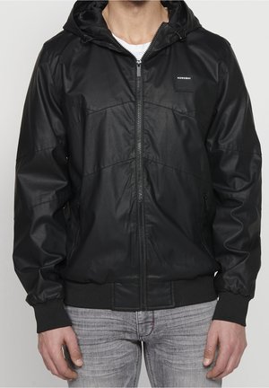 Mężczyzna noszący czarną kurtkę z kapturem na suwak, z żebrowanymi mankietami i dolnym ściągaczem, w połączeniu z jasnoszarymi dżinsami.