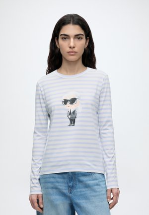 Γυναίκα με μακριά σκούρα μαλλιά φοράει ένα λευκό και γαλάζιο ριγωτό μακρυμάνικο πουκάμισο με μια καρτουνίστικη φιγούρα με γυαλιά ηλίου και μπλε τζιν.