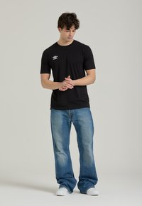 T-shirt nera a maniche corte con un logo sul lato sinistro del petto, abbinata a jeans in denim azzurro chiaro e sneaker bianche. Sfondo semplice.