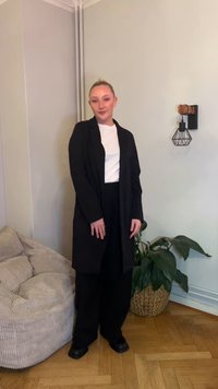 Cappotto lungo nero sopra una camicia bianca, abbinato a pantaloni neri a gamba larga e scarpe nere; ambientato in uno spazio interno minimalista con una pianta.