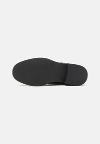 Semelle de chaussure noire avec une finition lisse et texturée. Forme arrondie, en caoutchouc, comportant une légère voûte et une imprimé de logo sur le côté.