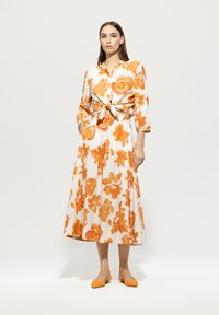 Blumenkleid in Weiß mit orangefarbenen Mustern, mit gebundenem Taillengürtel, langen Ärmeln und Maxilänge. Kombiniert mit orangen Ballerinas.