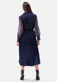 Navy blue pleated skirt with a fitted waistband, paired with a sheer long-sleeve blouse. Model wears black ankle boots.