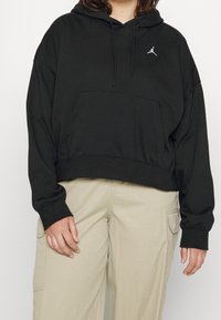 Sudadera negra corta con bolsillo frontal, que presenta un logo en el pecho. La tela es suave, con mangas largas y puños acanalados. Combinada con pantalones beige.