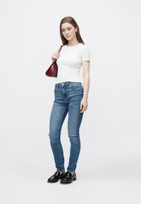 Jeans ajustados de mezclilla azul medio, combinados con una camiseta blanca ceñida. Lleva un pequeño bolso burdeos y usa mocasines negros.