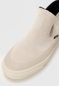 Beżowe zamszowe buty typu slip-on z elastycznymi panelami bocznymi, gumową podeszwą i teksturowanym noskiem. Posiada dyskretną metkę z logo blisko pięty.