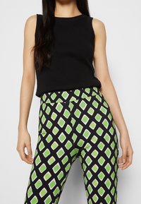 Kobieta ubrana w bezrękawnik w kolorze czarnym oraz wzorzyste spodnie z zielono-białymi diamentami na czarnym tle.