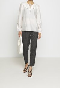 Blouse blanche légère et transparente avec col ruffle; pantalon noir à rayures; sandales noires à lanières; sac à main blanc.