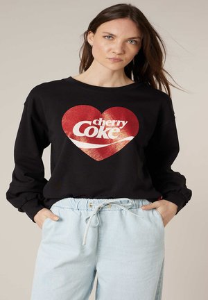 Femme aux longs cheveux bruns portant un sweat-shirt noir avec un cœur rouge et le texte "cherry Coke", associé à un pantalon bleu clair à cordon de serrage.