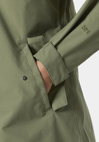 Αδιάβροχο μπουφάν σε ελιά πράσινο με λεία υφή, που διαθέτει πλαϊνή τσέπη με ορατό μανταλάκι και ραμμένη λεπτομέρεια στο μανίκι.