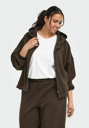 Femme aux cheveux foncés en queue de cheval, portant un t-shirt blanc, un sweat à capuche marron zippé et un pantalon de jogging marron assorti, souriante et regardant vers le bas.