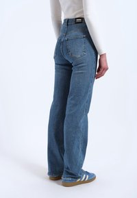 Personne portant un jean droit bleu et des baskets bleues à rayures blanches, debout de dos devant un fond blanc.