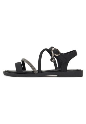 Sandalia plana negra con tres tiras delgadas, una con brillo, una correa con hebilla en el tobillo, y un pequeño colgante circular que cuelga de la hebilla.