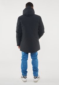 Giacca a vento nera lunga con cappuccio, caratterizzata da una texture liscia e un taglio dritto. Indossata con jeans azzurri chiari e sneakers nere.