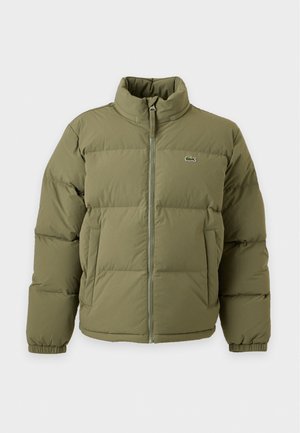 Un homme présente une doudoune vert olive, un polo bleu marine, une casquette et un pantalon beiges, ainsi que des baskets blanches, devant un fond de studio uni.