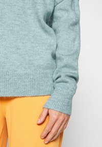 Světlý modrozelený pletený svetr s žebrovanými manžetami a lemem, s jemnou texturou. Kombinován se světlými žlutými kalhotami, které mají uvolněný střih.