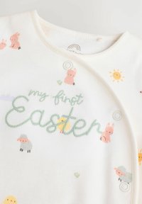 Hvid bomuldsromper med pastelfarvede påske-tema grafik, herunder kaniner, kyllinger og sol, med "min første påske" i grøn broderi.