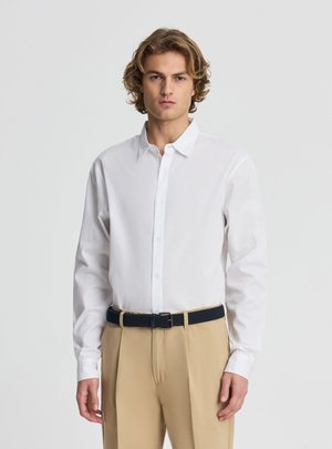 Uomo con capelli biondi ondulati che indossa una camicia bianca con bottoni, pantaloni beige e una cintura nera, in piedi davanti a uno sfondo chiaro e semplice.