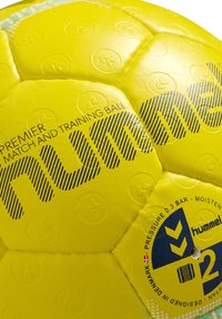 Gul träningsboll med texturerad yta; har svart logotyp och text, hexagonala paneler samt en tryckindikator på en hängetikett.