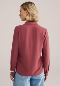 Camisa de manga larga con cuello en tono rosa apagado, con tela texturizada y puños abotonados, combinada con vaqueros de mezclilla azul claro.