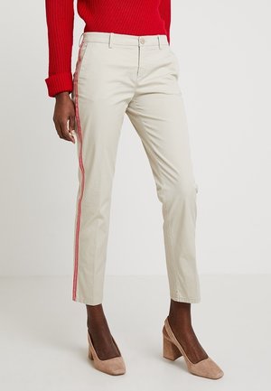 Mujer con pantalones ajustados beige con franjas laterales rojas, un suéter rojo y zapatos beige de tacón grueso, posando frente a un fondo liso.