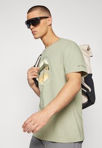 Lys grøn t-shirt med et cirkulært grafisk motiv, der viser bjerge og en solnedgang. Modellen har mørke solbriller på og bærer en rygsæk.