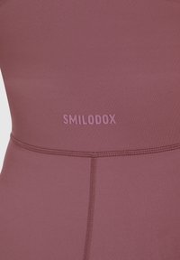 Bordeaux sporttop gemaakt van een gladde, rekbare stof met een opvallend roze "SMILODOX"-logo en subtiele naadtoespraken.