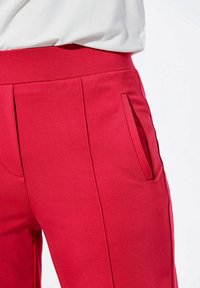 Eng anliegende rote Hose aus glattem Stoff, mit einer Seitentasche und dezenten Falten an den Hüften für Form und Detail.