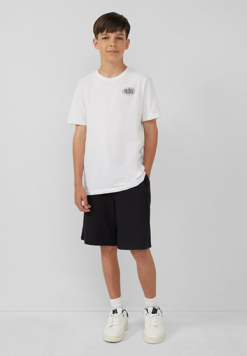 Witte katoenen t-shirt met een zwart logo, gecombineerd met zwarte shorts en witte sneakers met marineblauwe accenten. Eenvoudige, casual stijl.