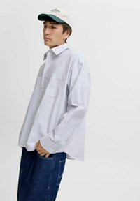 Ung mand iført en lysstribet skjorte med knapper, mørke jeans og en hvid og grøn kasket, poserer mod en simpel lys baggrund.