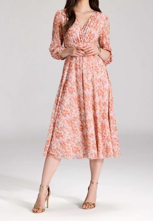 Femme portant une robe midi fleurie rose à manches trois-quarts et des sandales à talons hauts dorées, debout, les mains jointes.