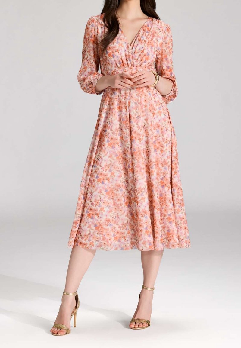 Femme portant une robe midi fleurie rose à manches trois-quarts et des sandales à talons hauts dorées, debout, les mains jointes.