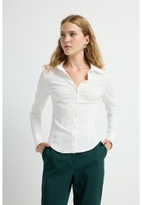 Camicia bianca a maniche lunghe con bottone frontale, colletto rigido e tessuto strutturato; abbinata a pantaloni verde scuro.