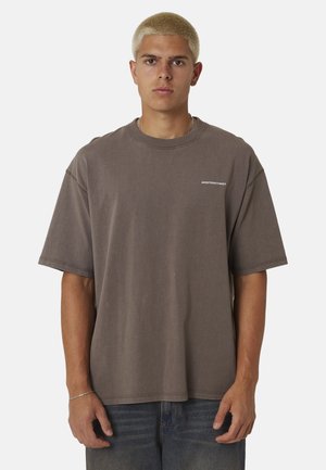 Jeune homme aux cheveux blonds courts, portant un t-shirt marron ample et un jean bleu délavé, debout devant un fond uni.