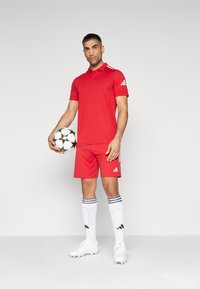 Rode polo met korte mouwen en shorts met witte adidas-accenten. Witte sportsokken met zwarte strepen en witte voetbalschoenen. Houdt een zwart-wit voetbאל.