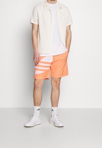 Valkoinen lyhythihainen paita ja valkoinen t-paita yhdistettynä oransseihin uima-shortseihin, joissa on valkoiset vinoraitaiset kuviot. Valkoiset lenkkarit ja valkoiset sukat.