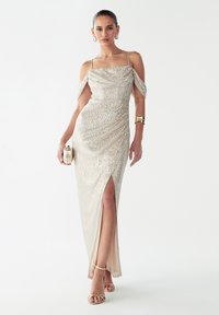 Večerna obleka s perlicami v svetlo zlati barvi, z odkrito ramenskiimi rokavi, stranskim razporkom in oprijeto silhueto. Kombinirana z zlatimi sandali s trakovi.