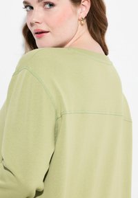 Camicia a maniche lunghe verde chiaro realizzata in tessuto morbido. Presenta una vestibilità rilassata e sottili dettagli di cucitura lungo il girocollo e le spalle.