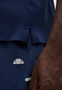Gros plan sur une main à la peau foncée à côté d'un short de sport bleu marine avec un petit logo blanc et un ourlet de chemise bleu marine texturé.