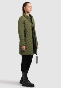 Lunga giacca trapuntata verde oliva con colletto alto, tasche laterali con zip e accessori neri, abbinata a stivali neri alti fino al ginocchio con lacci.