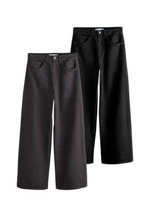 Person iført sorte jeans, grå skjorte og beige jakke med hænderne afslappet i lommerne.