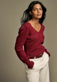 Pullover bordeaux lavorato a maglia con scollo a V, texture a costine e maniche a sbuffo, abbinato a pantaloni color crema e una cintura marrone.