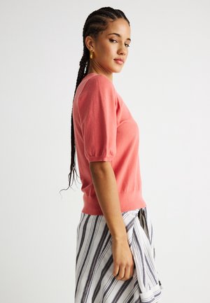 Joven con el cabello trenzado, llevando pendientes de aro dorados, una blusa coral de manga corta y una falda de rayas, sobre un fondo blanco liso.