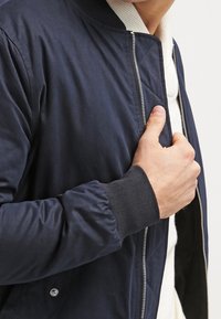 Granatowa kurtka bomberka o gładkiej fakturze, z prążkowanymi mankietami i srebrnym zamkiem błyskawicznym. Wewnętrzna warstwa posiada jasny, dzianinowy kołnierz.